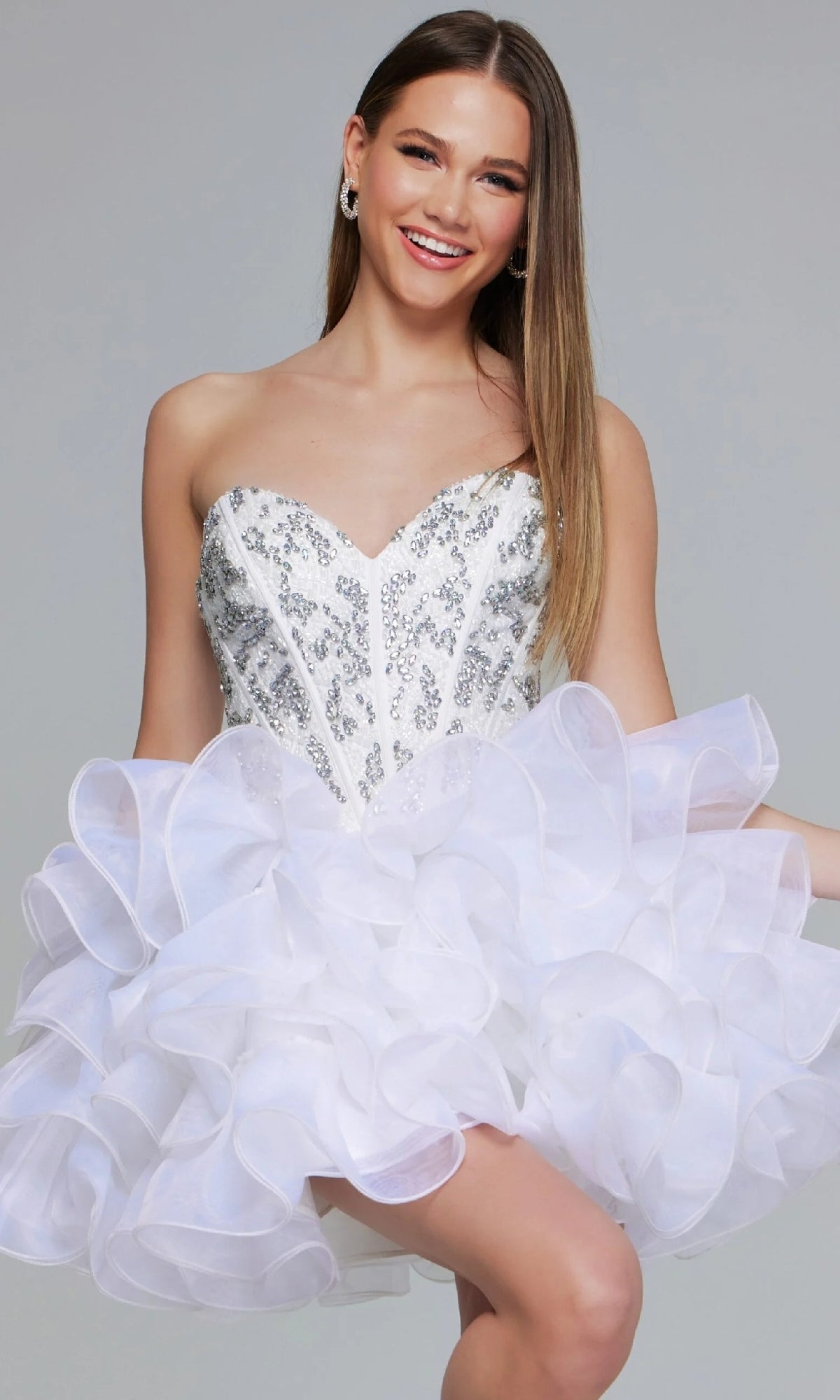 A woman smiles against a plain background, wearing the Jovani 39932 Short Homecoming Dressโa strapless, white sequin dress with a sweetheart neckline and a ruffled, layered skirt.